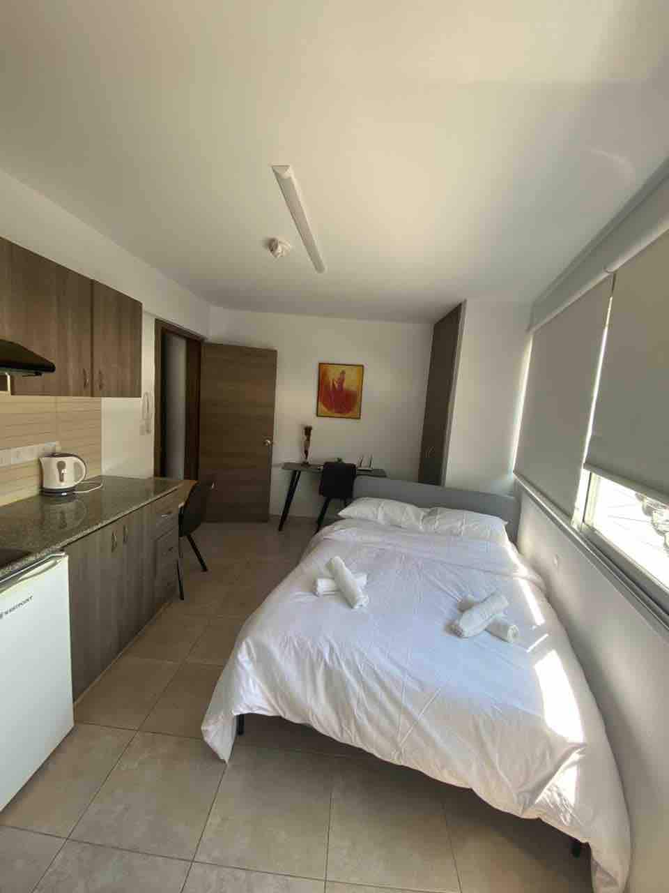 A compact studio layout is observed, featuring a spacious bed with freshly arranged linens. A small kitchenette is present, equipped with essential appliances. Natural light enters through window blinds, creating a bright atmosphere. A wooden dining chair and a small table complete the furnishings.
