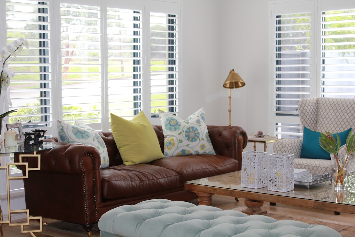 The living area features a combination of a brown leather sofa and a patterned armchair, both positioned near large windows adorned with shutters. A coffee table with a glass top rests on a tufted ottoman, complemented by decorative pillows and a side lamp.