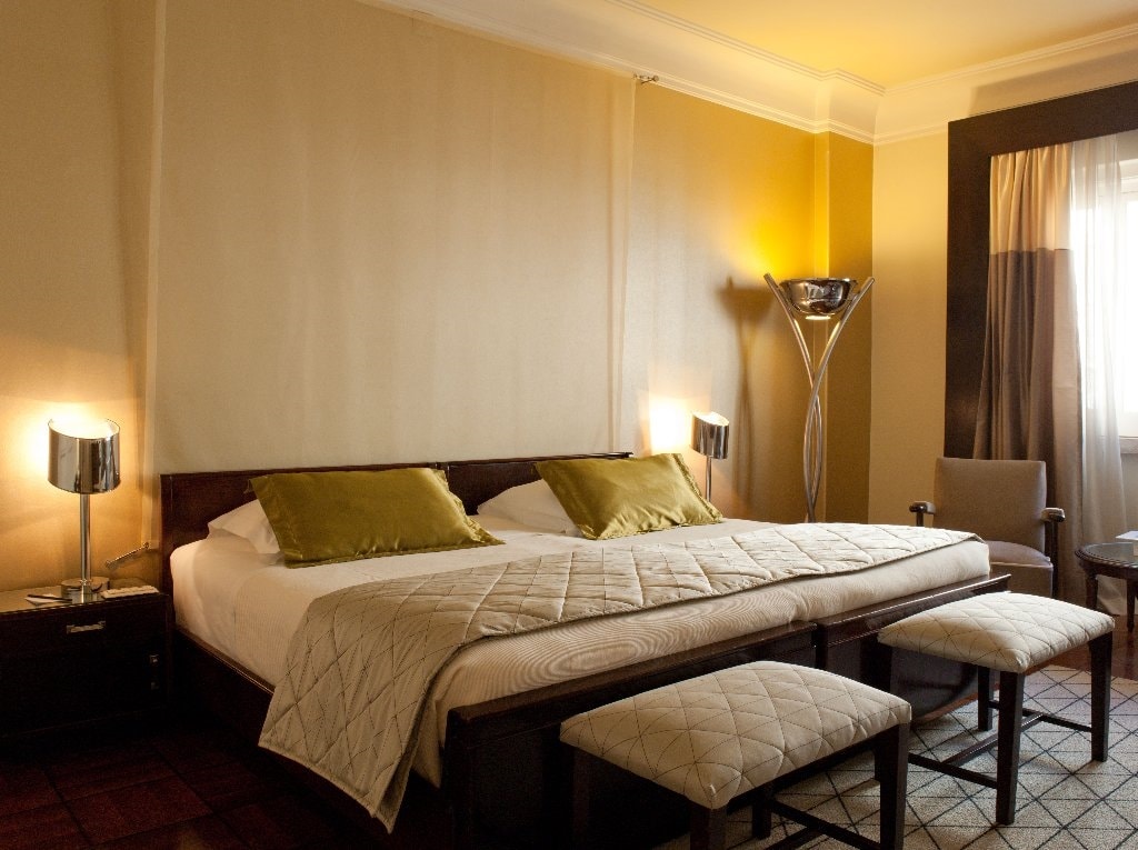 A spacious room features a double bed topped with two green cushions. Soft lighting from floor lamps creates a cozy ambiance. A comfortable armchair and a round side table are positioned nearby, while a window allows natural light to enter, complemented by a light curtain.