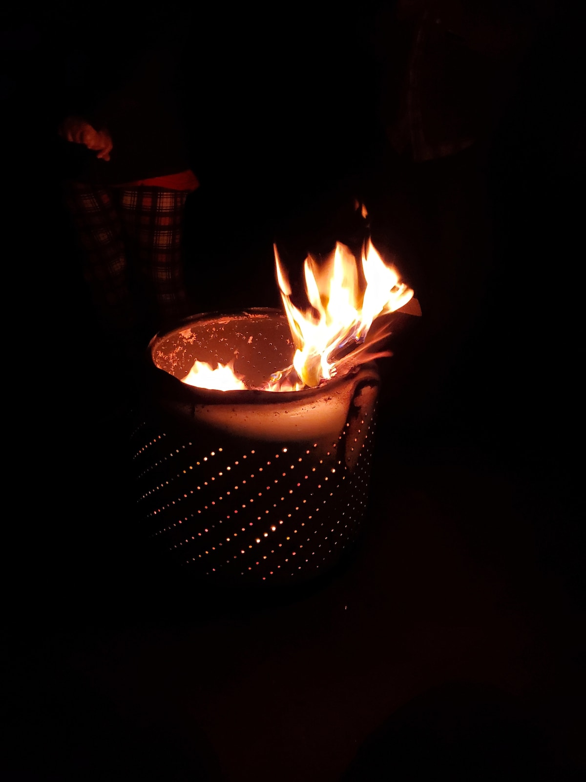 A metallic fire pit is shown, emitting bright flames against a dark background. The surface of the pit features small perforations, allowing light to escape. The glow from the fire creates a warm ambiance, visible in the nearby shadows.