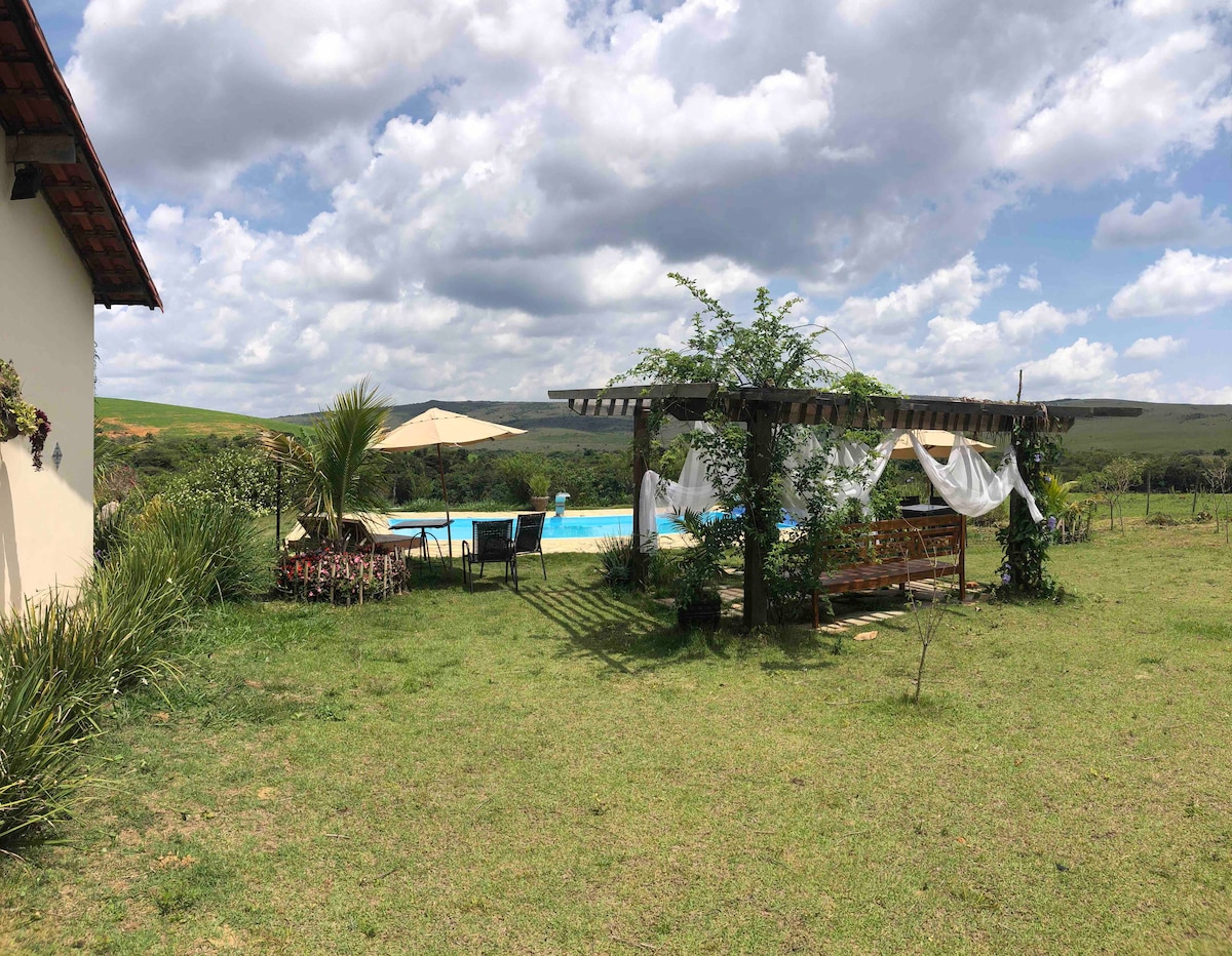 The outdoor area displays a spacious lawn with a pergola draped in white fabric, providing shaded seating. A clear blue pool is visible in the background, surrounded by lush greenery and an umbrella. The sky features scattered clouds, contributing to a serene natural setting.