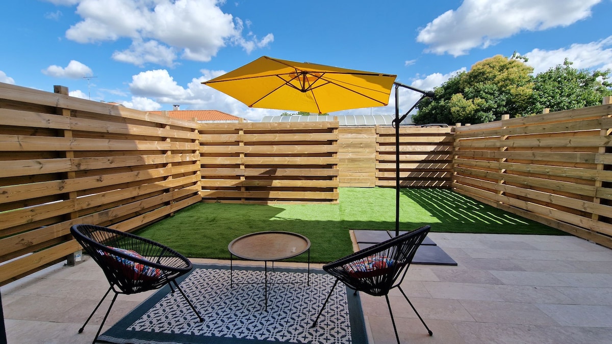 A private outdoor space is featured, showcasing a vibrant yellow umbrella providing shade over a small table and two black lounge chairs. The area is surrounded by wooden fencing, complemented by artificial grass, creating a serene and inviting atmosphere.