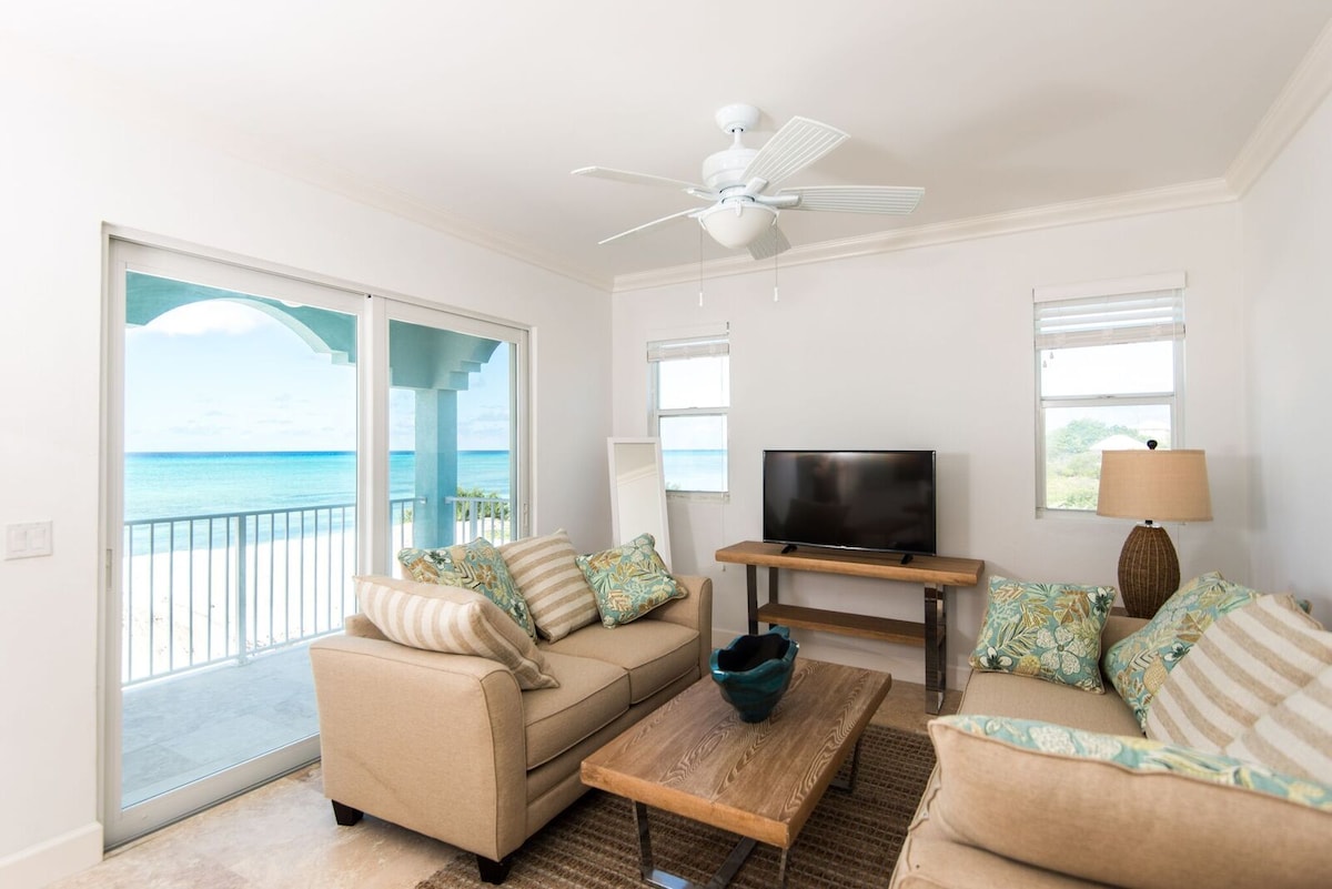 A comfortable living area is featured, showcasing a light-colored sofa adorned with decorative pillows. A wooden coffee table is positioned in front, with a blue bowl atop it. A flat-screen TV is mounted on a wooden stand, and large sliding glass doors provide a view of the ocean.