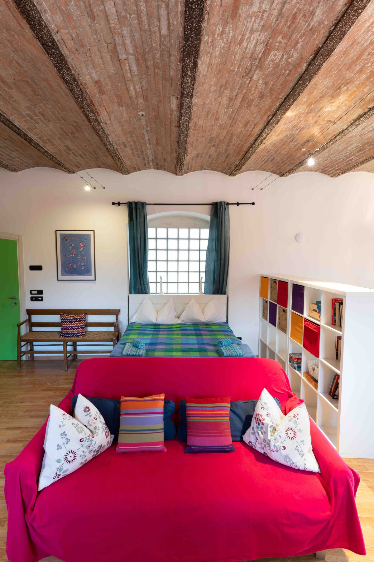 An open space features a vibrant red sofa adorned with decorative pillows, positioned in front of a bed covered with a colorful blanket. Exposed brick beams span the ceiling, and a window draped with blue curtains provides natural light. A bookshelf and a small dining area complete the setting.
