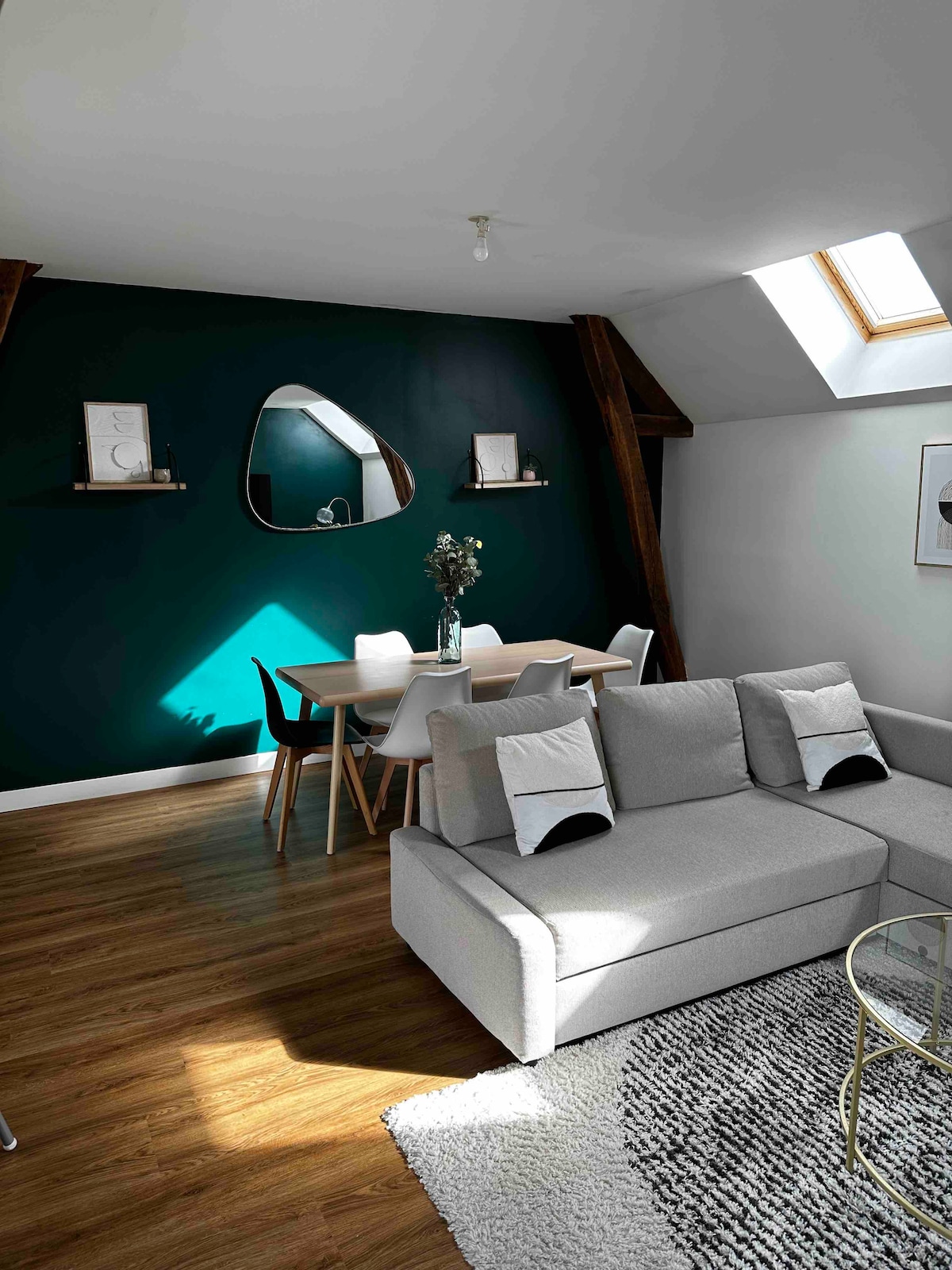 A spacious living area features a modern gray sectional sofa and a stylish glass coffee table. A dining table with white chairs is positioned against a deep green accent wall. Natural light enters through a skylight, highlighting the wooden beams above.