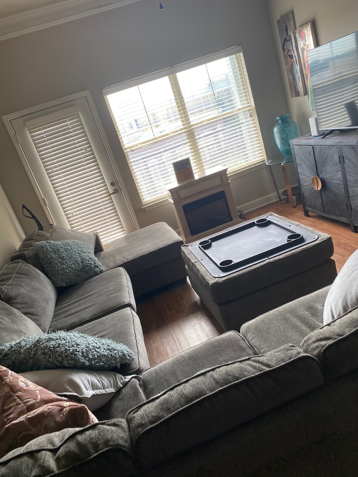 A cozy living area features a sectional sofa adorned with decorative pillows, complemented by a coffee table with built-in cup holders. Natural light streams through a large window adorned with blinds. A television sits on a cabinet, and a small fireplace adds warmth to the space.