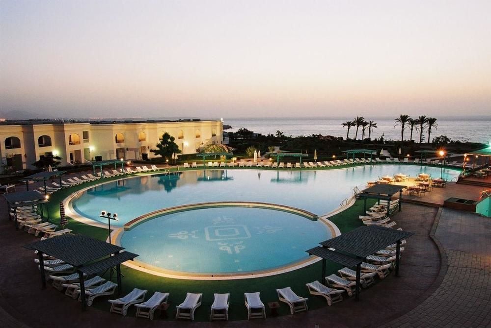 An expansive swimming pool is framed by neatly arranged lounge chairs, with cozy seating areas visible at the poolside. The tranquil water reflects the evening sky, while palm trees line the horizon, providing a sense of relaxation and harmony.