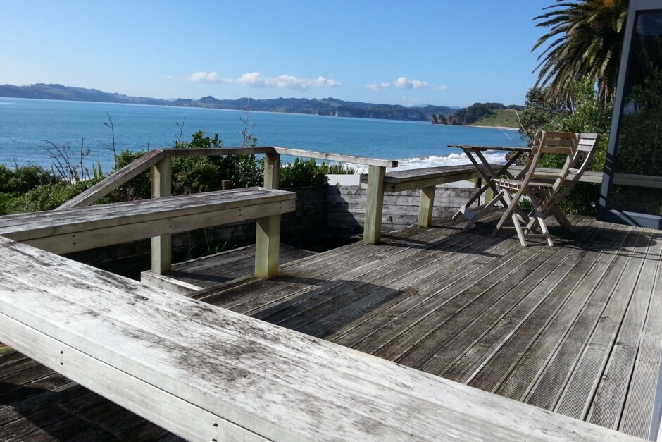 A spacious wooden deck overlooks Cook's Beach, offering panoramic views of the water and nearby cliffs. Two wooden foldable chairs are positioned invitingly, with the tranquil sea gently lapping at the shore in the background. Lush greenery frames the scene, enhancing the serene atmosphere.