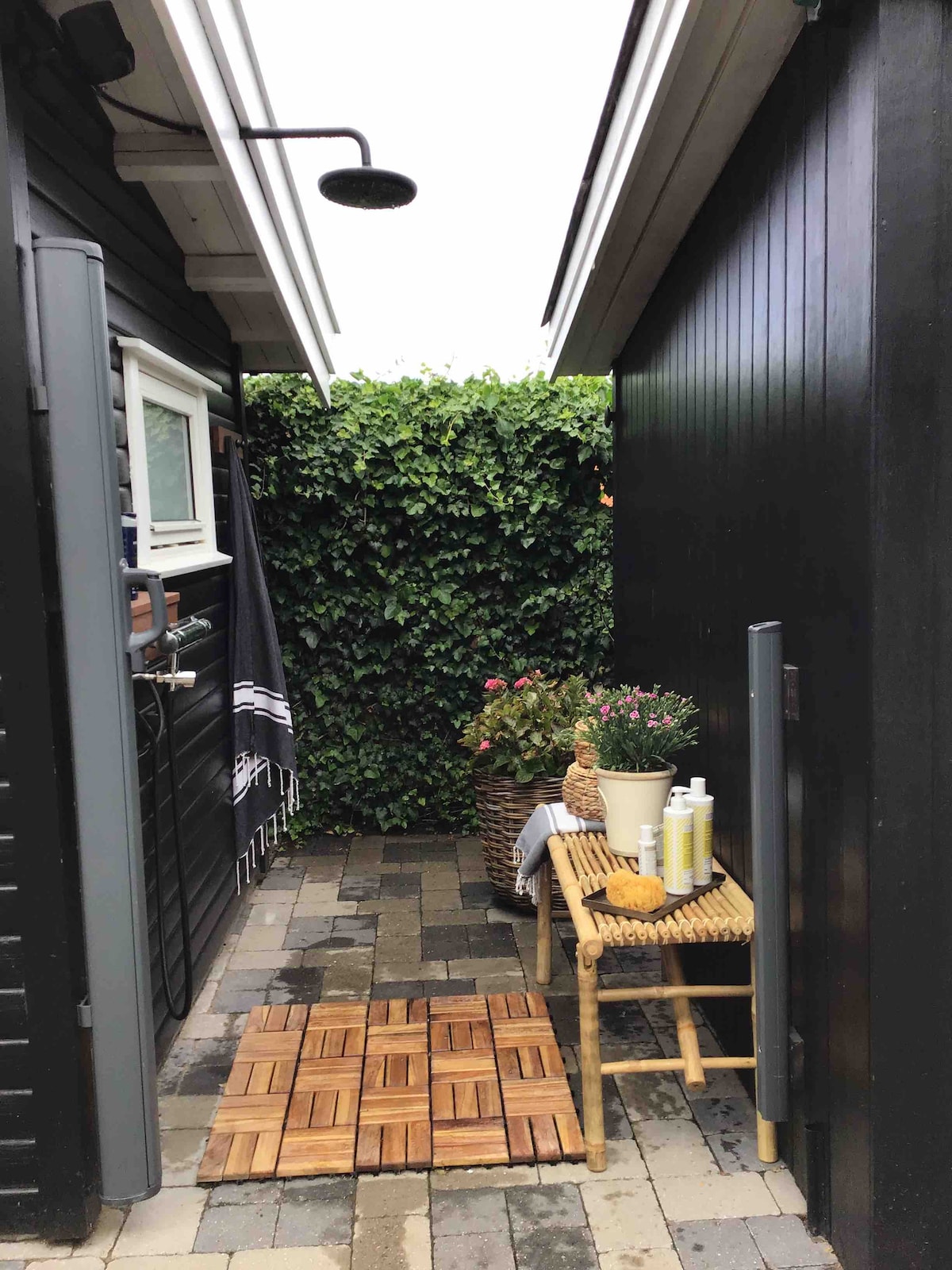 An outdoor shower area is positioned between two walls, featuring a rainfall showerhead. A wooden mat leads to a small wicker table adorned with toiletries, surrounded by a lush green hedge and potted plants, creating a private and serene space.