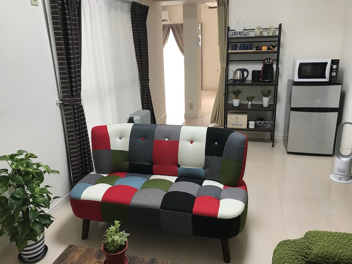 A colorful patchwork sofa with a mix of red, green, and white fabric sits in a bright room. A small potted plant rests on the floor nearby, while a bookshelf and kitchenette with a microwave and coffee maker are visible in the background.