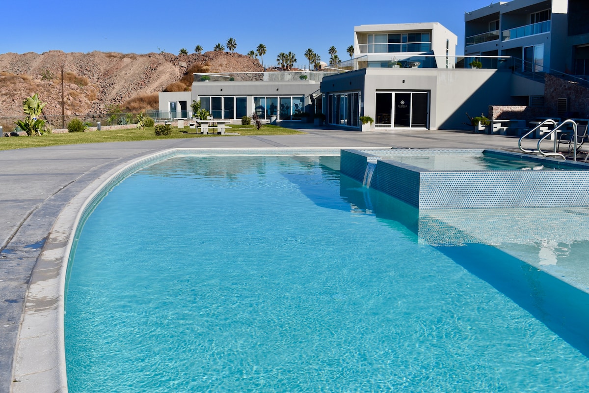 A spacious outdoor pool is featured, showcasing clear blue water with a shallow area designed for relaxation. Surrounding the pool, sun loungers are arranged, and the contemporary building is visible in the background, with large glass doors allowing for natural light.