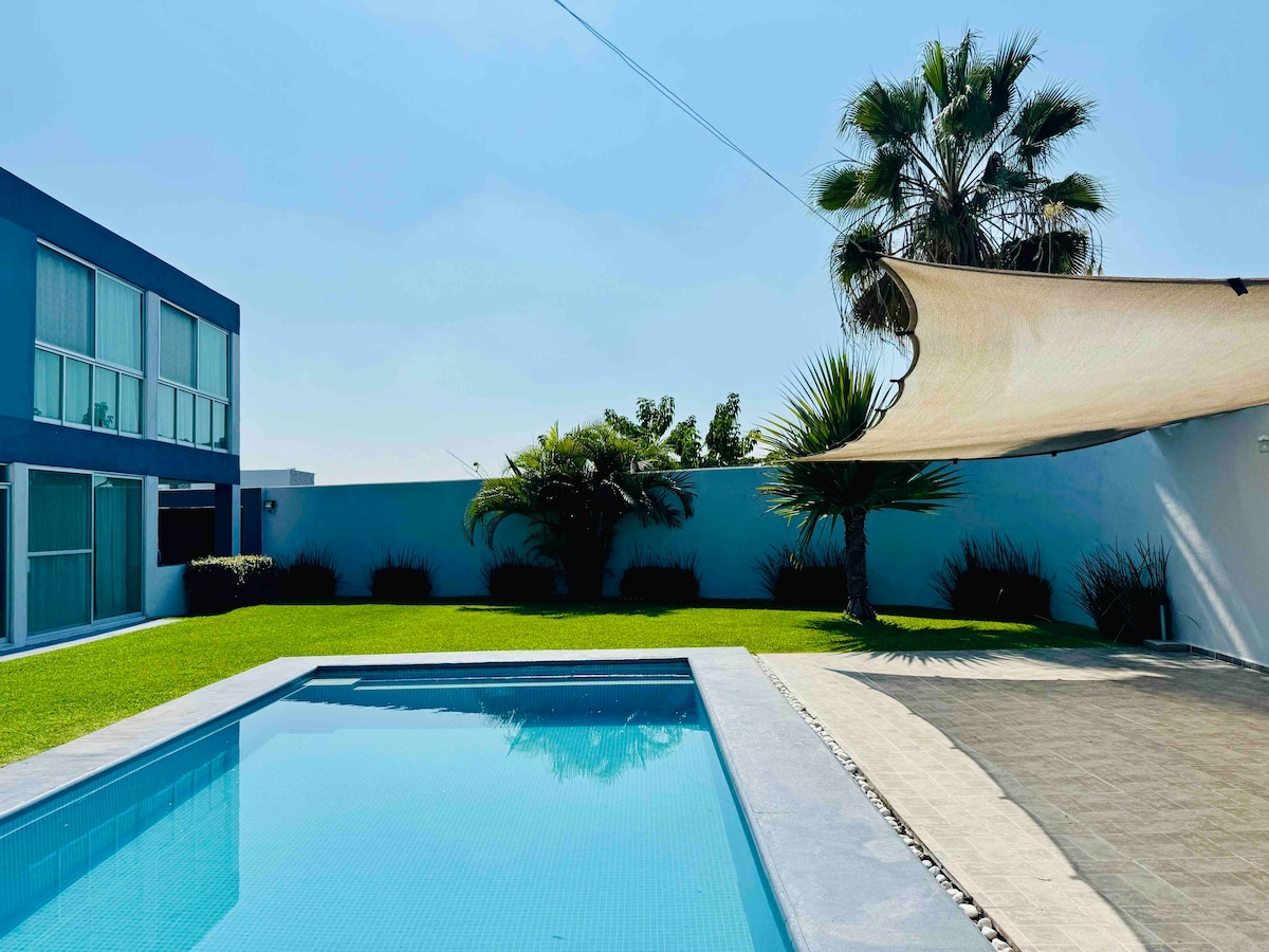 A serene outdoor space features a swimming pool surrounded by lush green grass. A shaded area with a sail extends over the pool, while palm trees and other greenery enhance the tranquil setting. The clear blue sky completes the inviting atmosphere.