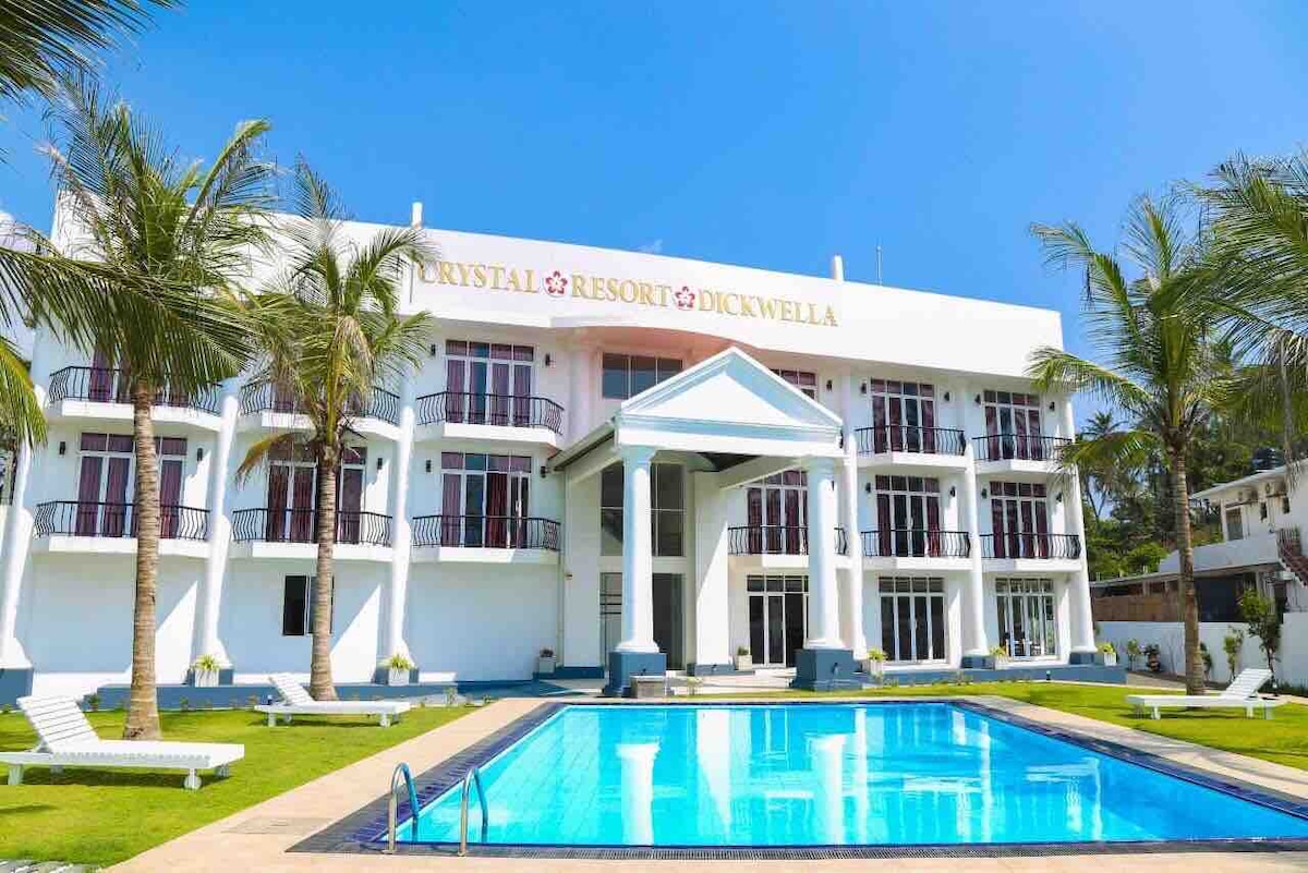A modern resort building features a symmetrical façade with multiple balcony spaces and large glass windows. A rectangular pool is centered in front, reflecting the clear blue sky, while palm trees surround the landscaped grounds, contributing to the tropical atmosphere.