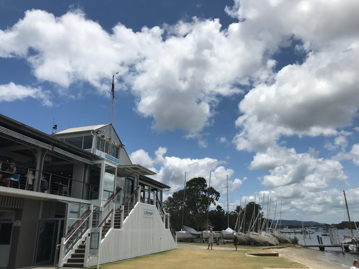 The image depicts a waterfront building with a staircase leading to its entrance. A grassy area is visible in the foreground, with several sailboats moored nearby. Fluffy clouds scatter across the blue sky, creating a serene backdrop for the scene.
