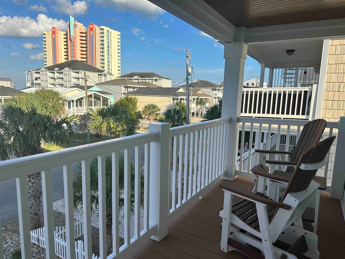 A spacious balcony is showcased with two wooden rocking chairs, providing a view of nearby buildings and palm trees. The railing features white pickets, while the flooring is crafted from wood. The sky is bright with clouds, enhancing the outdoor setting.