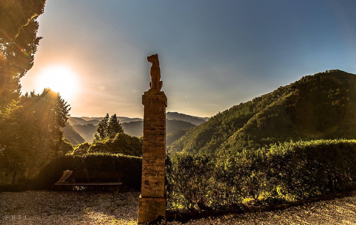 A stone pillar featuring a carved figure stands prominently against a backdrop of rolling hills. The sun rises over the horizon, casting a warm glow across the landscape, while lush greenery blankets the mountainside.