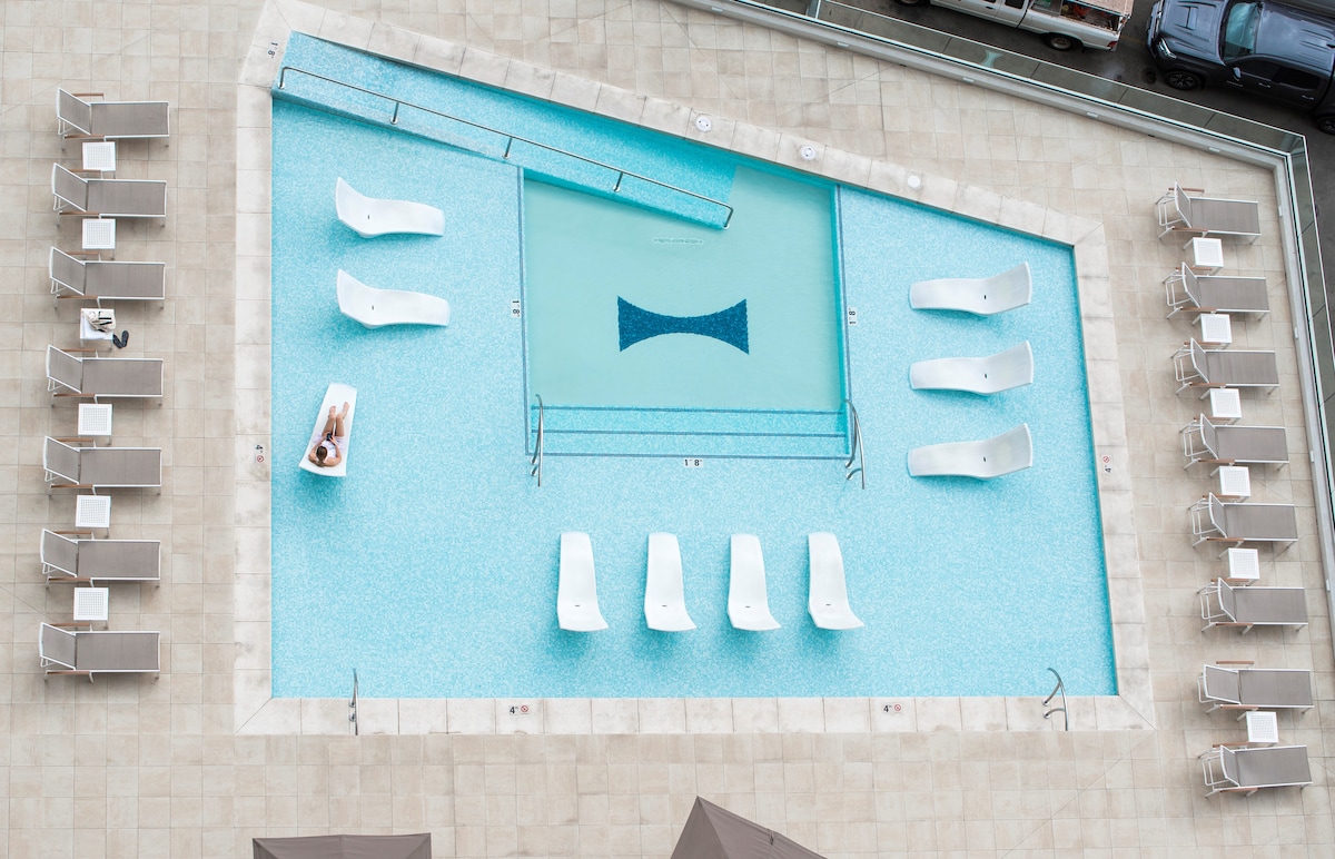 An expansive pool area is highlighted by a unique geometric design. Multiple loungers surround the pool, providing ample space for relaxation. The pool's water glistens in a vibrant blue hue, inviting guests to enjoy the refreshing atmosphere.