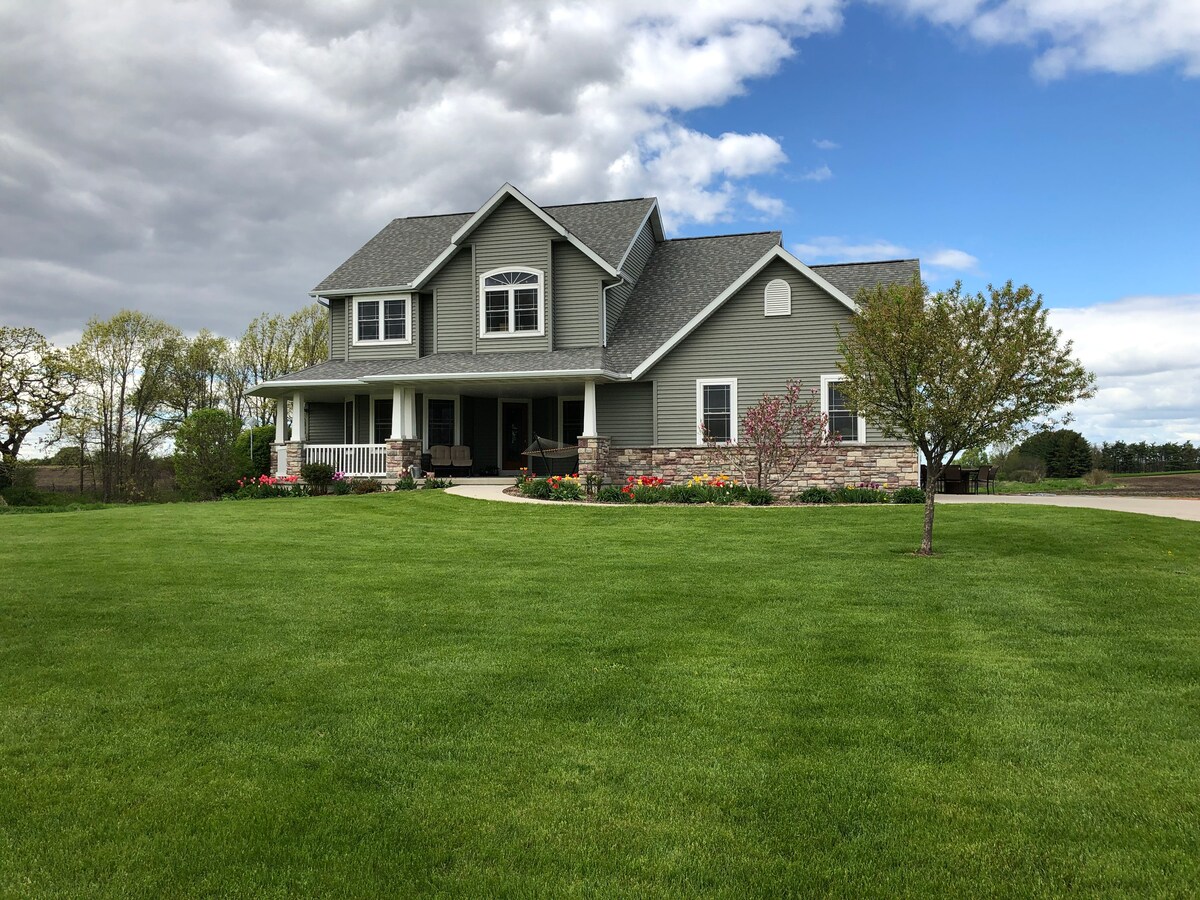 The two-story house features a covered porch with seating, surrounded by blooming flower beds and lush green grass. Large windows are apparent on both levels, and the exterior is finished in muted gray with stone accents. An expansive lawn leads to a paved driveway.