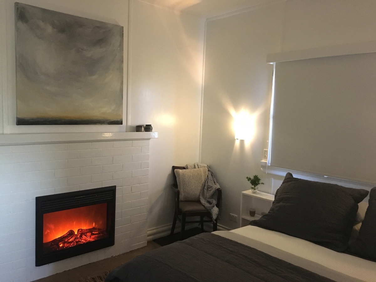 A cozy bedroom features a spacious bed with grey linens, complemented by a minimalist chair positioned nearby. A modern fireplace, set within a white-brick wall, provides warmth, while soft lighting enhances the room's comfortable ambiance. Artwork hangs above the fireplace, adding a touch of creativity.