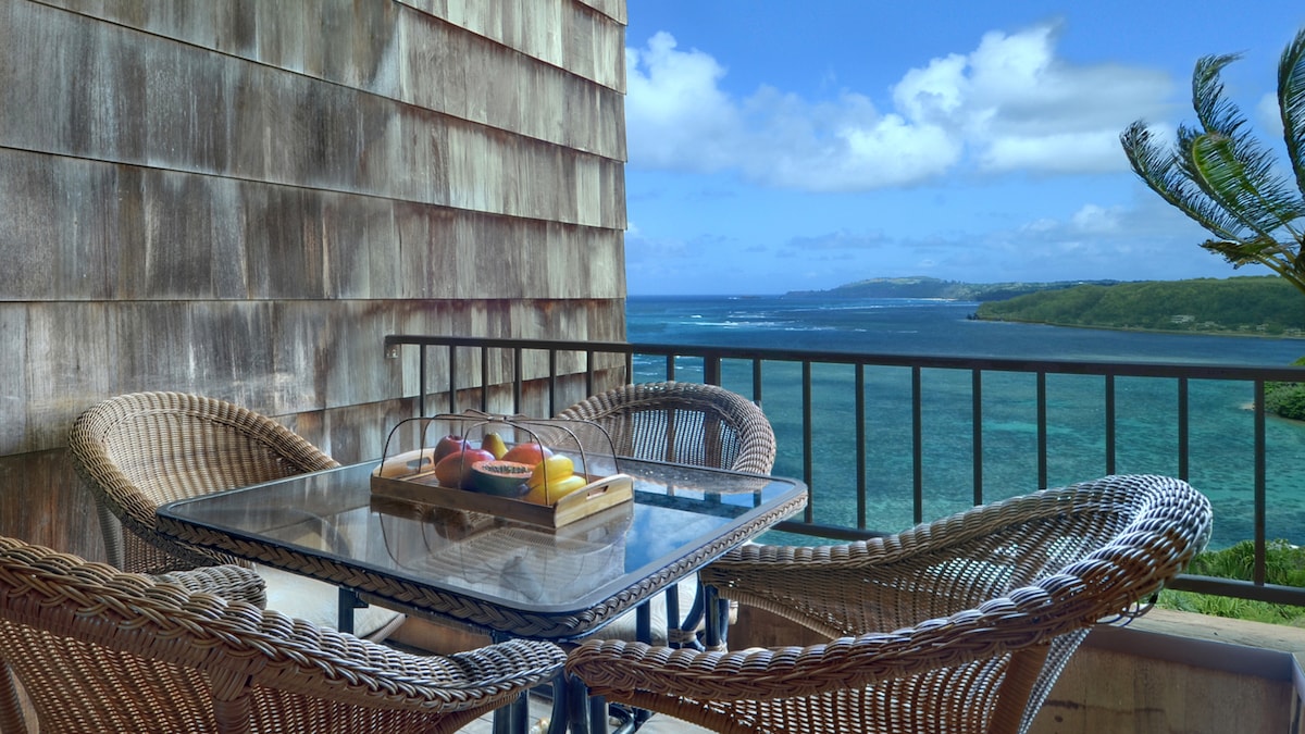 A private covered lanai features a table set with a bowl of fresh fruit and four woven chairs. The ocean view reveals calm waters with distant green hills under a partly cloudy sky, creating a serene atmosphere.