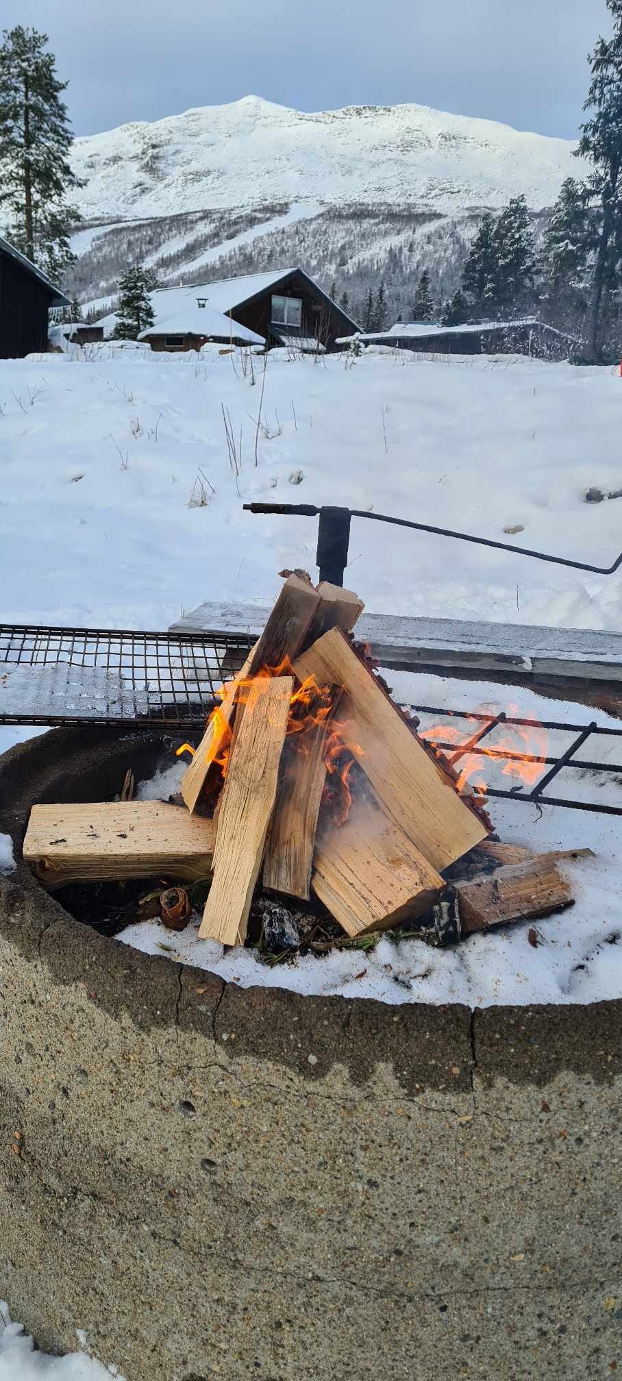 A fire pit is filled with neatly stacked firewood, flames visible as they flicker and dance upward. Snow covers the surrounding ground, and the backdrop includes distant cabins and a mountain landscape under a gray sky.
