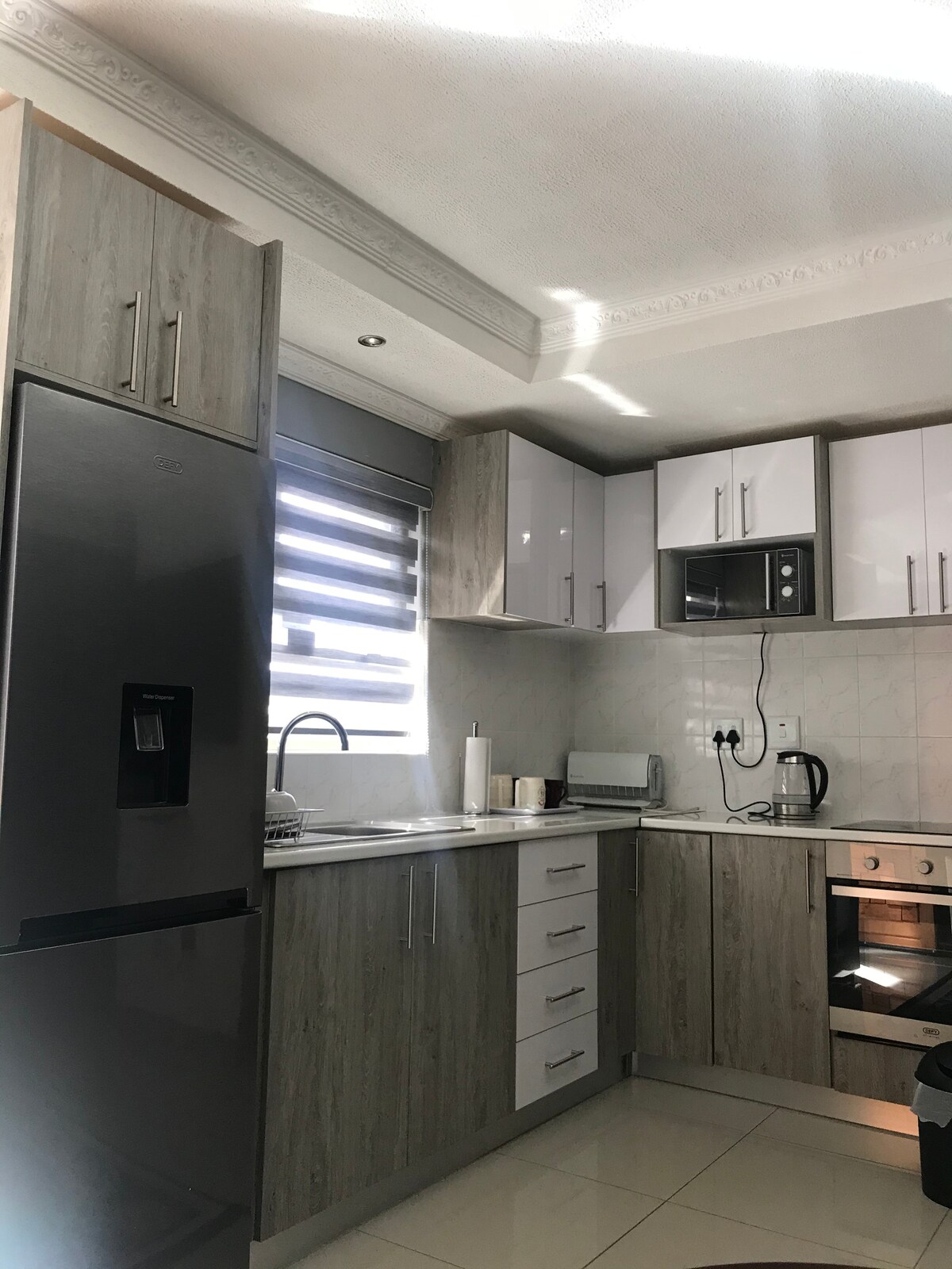 A modern kitchen features sleek cabinetry in neutral tones with contrasting white accents. Stainless steel appliances, including a refrigerator and oven, are visible. Natural light enters through the window, highlighting the counter space and sink area, which are equipped with essential cooking tools.