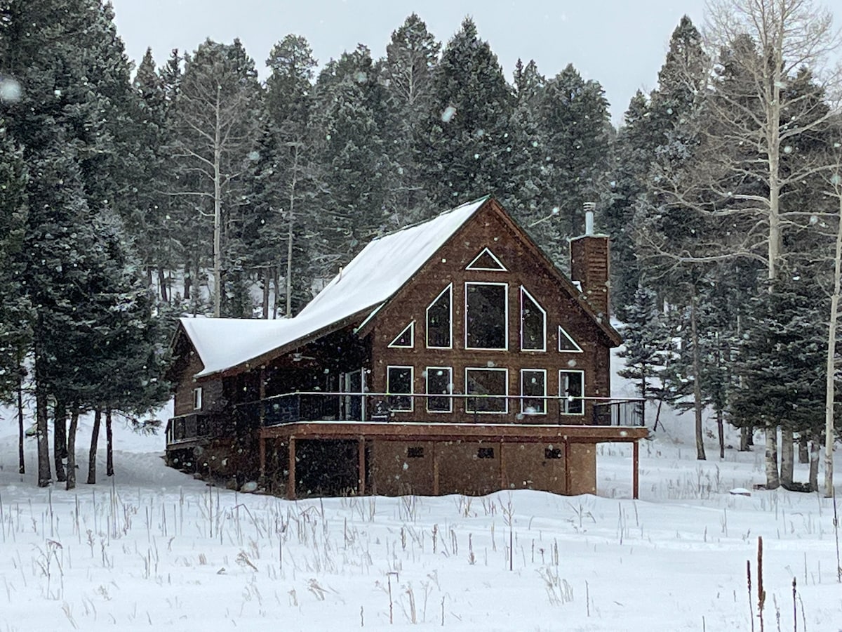 A spacious wooden home is situated among tall trees, with a snow-covered landscape surrounding it. The sloped roof features multiple gables, and large windows allow for natural light to fill the interiors. A deck extends from the front, providing an outdoor space.