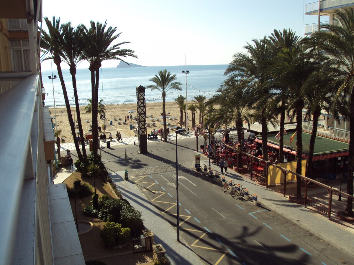A view from the balcony showcases the sandy beach and calm sea, with palm trees lining the shore. An empty street is visible, leading to nearby cafes and leisure activities. Sunlight brightens the scene, indicating a warm, inviting atmosphere along the promenade.