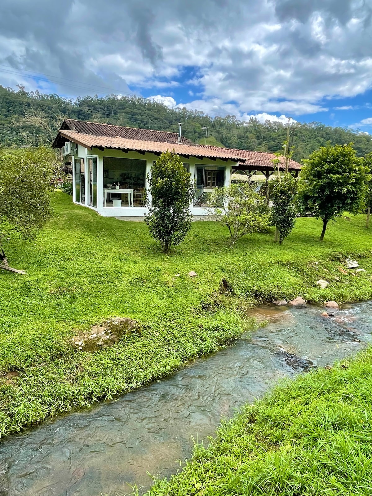 A charming house is nestled among lush greenery, with a stream flowing nearby. The structure features large windows, allowing for scenic views of the surrounding nature. The landscape includes well-maintained grass and ornamental trees, enhancing the peaceful outdoor setting.