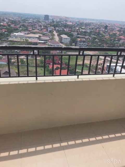 A spacious balcony is seen, featuring a black railing that overlooks a vast urban landscape. The view includes rooftops of nearby buildings and distant greenery, providing a sense of openness and connection to the surroundings.