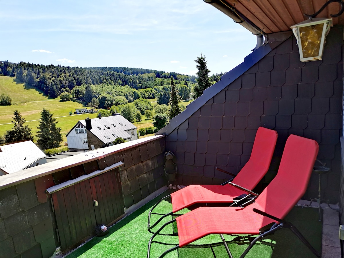 A private balcony is shown featuring two red lounge chairs positioned for relaxation. The scenic view of rolling green hills and wooded areas extends in the background, providing a calm outdoor atmosphere. The balcony is designed for exclusive use, emphasizing a sense of privacy.