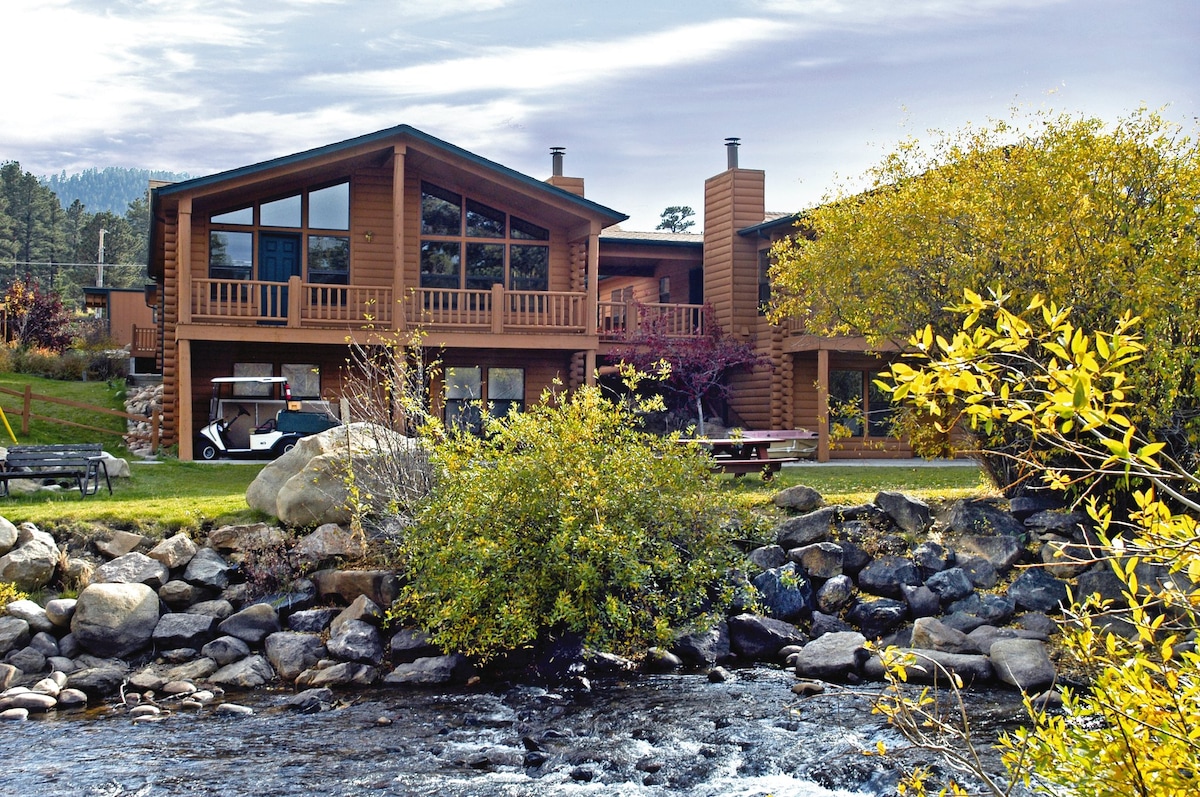 A multi-story wooden building features large windows that provide views of the surrounding landscape. A rocky riverbank is visible in the foreground, accompanied by vibrant yellow foliage. Outdoor seating can be seen near the structure, integrating with the natural setting.
