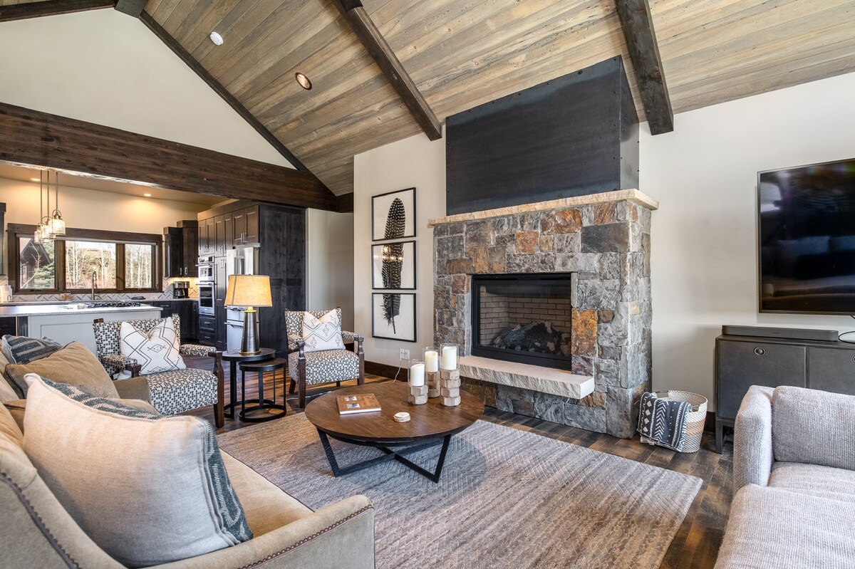 A spacious great room features vaulted wooden ceilings and a stone fireplace as a focal point. Comfortable seating arrangements surround a round coffee table, which holds decorative items. Soft lighting from a nearby lamp enhances the cozy ambiance, with a view into the kitchen area visible in the background.