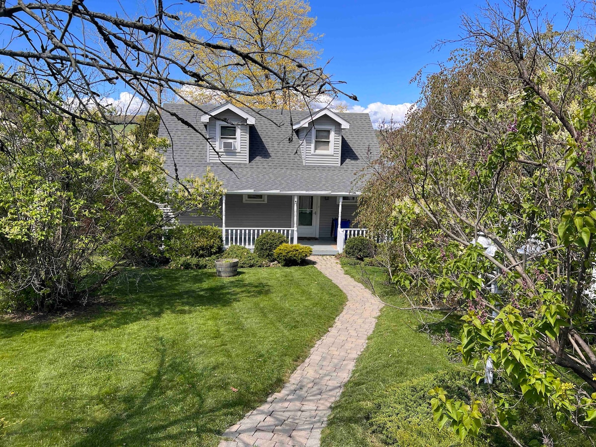 A charming cottage is framed by well-maintained gardens and lush green grass. A curving stone pathway leads to the front porch, enhancing the inviting entrance. The structure features a gabled roof and symmetrical windows, complementing the surrounding flora.