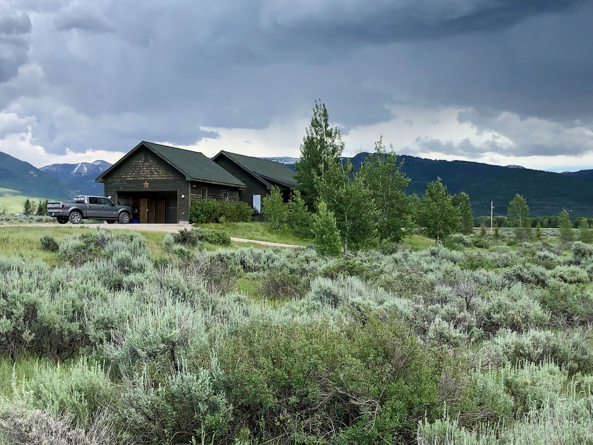 A charming vacation home is set amidst expansive sagebrush, surrounded by greenery. The structure features a dark exterior and a peaked roof, while a vehicle is parked in the gravel driveway. The backdrop displays a stunning view of distant mountains under a cloudy sky.