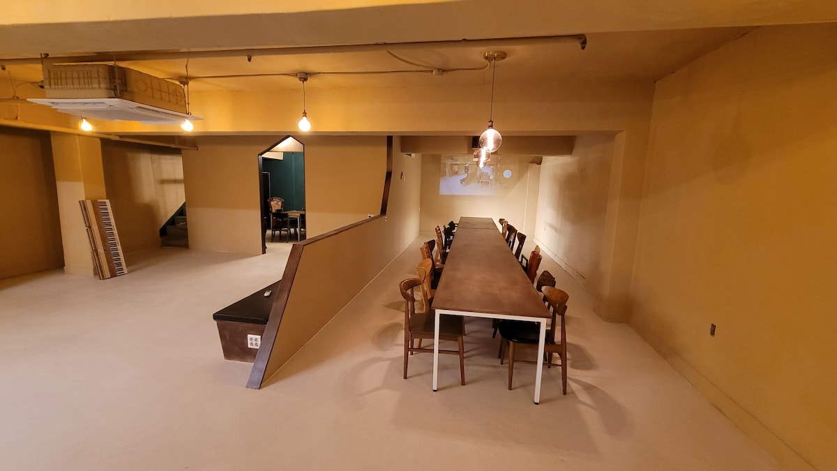 An expansive gathering space showcases a large table designed to accommodate multiple guests. Surrounding seating includes wooden chairs, complementing the minimalistic decor. Soft lighting from overhead fixtures enhances the spacious environment, with a wall-mounted screen visible at the far end.