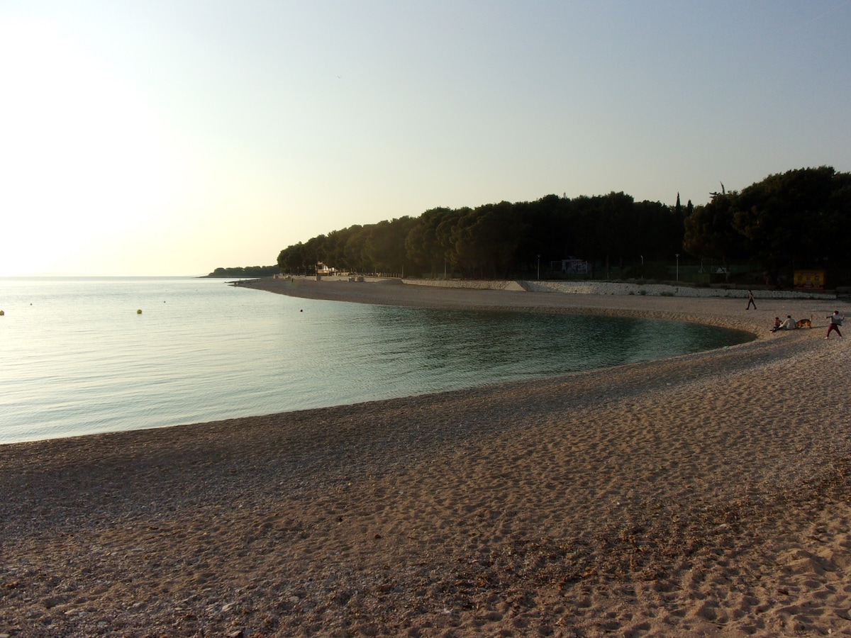 A calm beach scene is presented with gently lapping waves on a sandy shore. Lush greenery lines the coastline, while visitors can be seen enjoying the area. The horizon reflects the warm colors of sunset, creating a serene atmosphere.