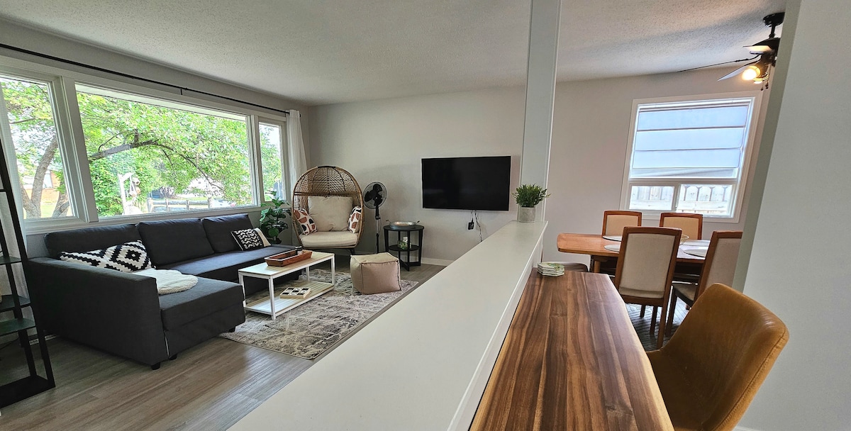 A bright living area is visible, featuring a comfortable dark sofa with decorative cushions and a large window offering natural light. A television is mounted on the wall opposite a hanging chair. A dining table for six is positioned in the background, with a view of the kitchen.