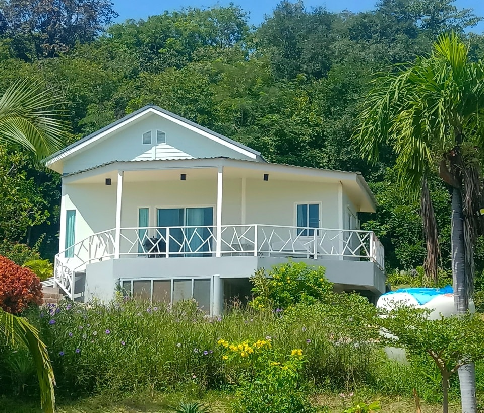 A modern building features a spacious deck with a railing, surrounded by vibrant greenery. Large windows provide natural light, while tropical plants enhance the outdoor area. The structure is set against a lush hillside, showcasing a harmonious blend with the natural environment.