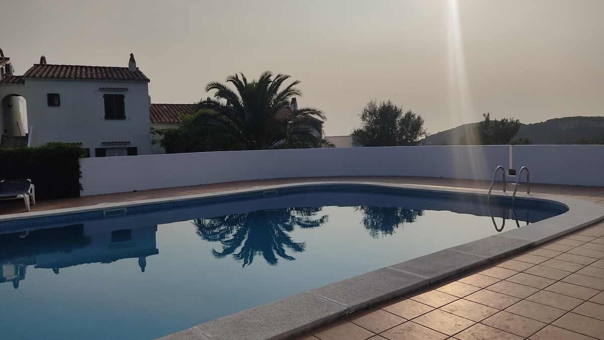 The image captures a tranquil swimming pool with clear water reflecting the sun and surrounding landscape. A tiled deck extends around the pool, framed by lush greenery and palm trees. The serene atmosphere suggests an inviting space for relaxation and leisure.