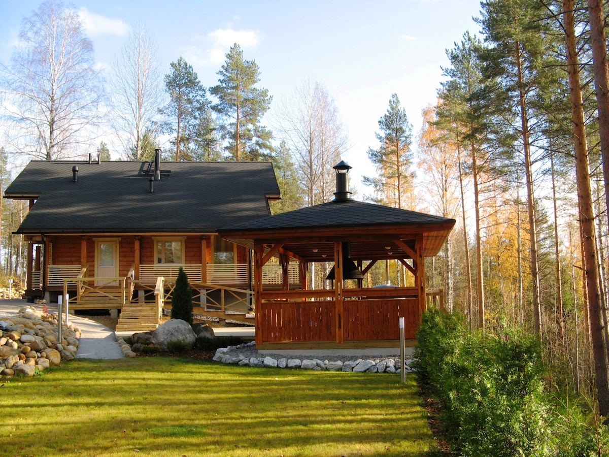 A modern wooden house is accompanied by a spacious gazebo featuring a chimney. Surrounded by lush green grass and trees, the scene offers a peaceful outdoor setting, ideal for relaxation. A clear pathway leads to the entrance, enhancing accessibility.