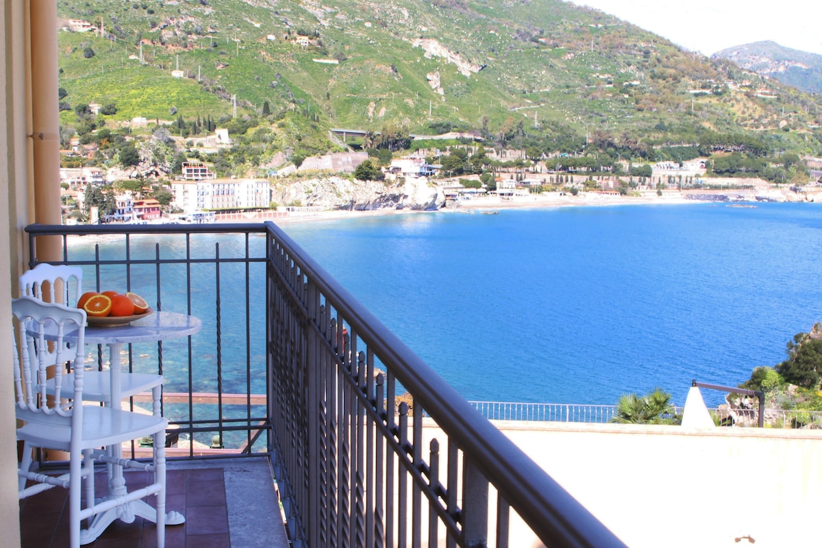 A balcony with outdoor furniture offers a clear view of the blue sea and surrounding hills. A small round table set with fresh fruit is positioned next to white chairs, inviting relaxation while enjoying the picturesque scenery.