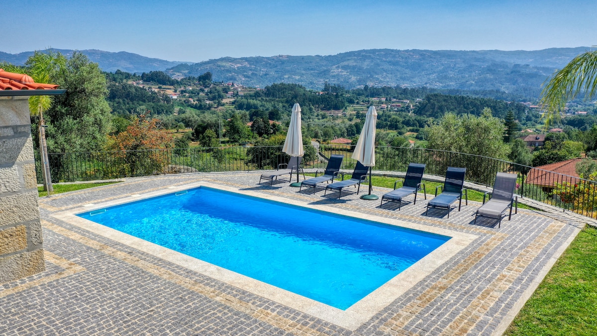 An inviting outdoor pool is surrounded by a stone patio, featuring several lounge chairs and two umbrellas for shade. Lush green hills and distant mountains are visible in the background, creating a serene setting for relaxation.