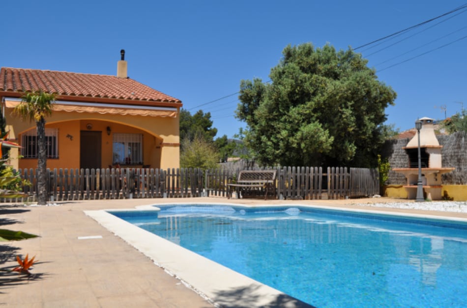 A single-story house is depicted with a red-tiled roof, surrounded by trees and greenery. A blue swimming pool occupies the foreground, bordered by tiled decking. A wooden bench sits near the pool, while a barbecue area is visible at the far end of the outdoor space.