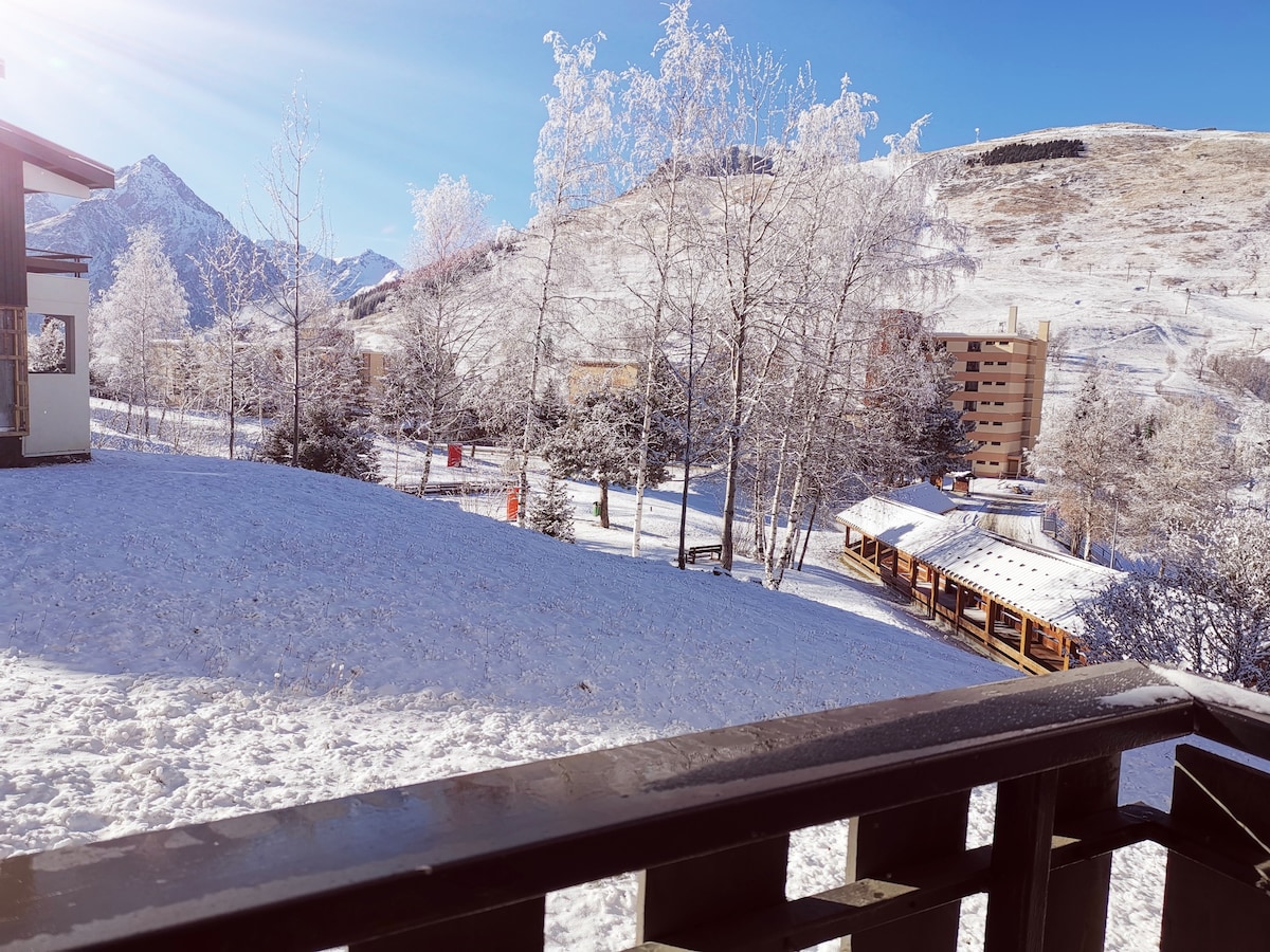 A winter landscape is displayed, showcasing a snow-covered field and trees with frost glistening in the sunlight. Mountains rise in the background, and colorful buildings can be seen nestled among the trees. A wooden structure offers shade in the distance.