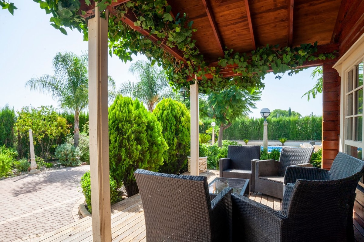 A shaded outdoor patio features a wooden ceiling and is furnished with dark wicker seating. Lush greenery surrounds the area, creating a private space, while a pathway leads away from the seating towards the garden. Sunlight filters through the leaves, casting gentle shadows.