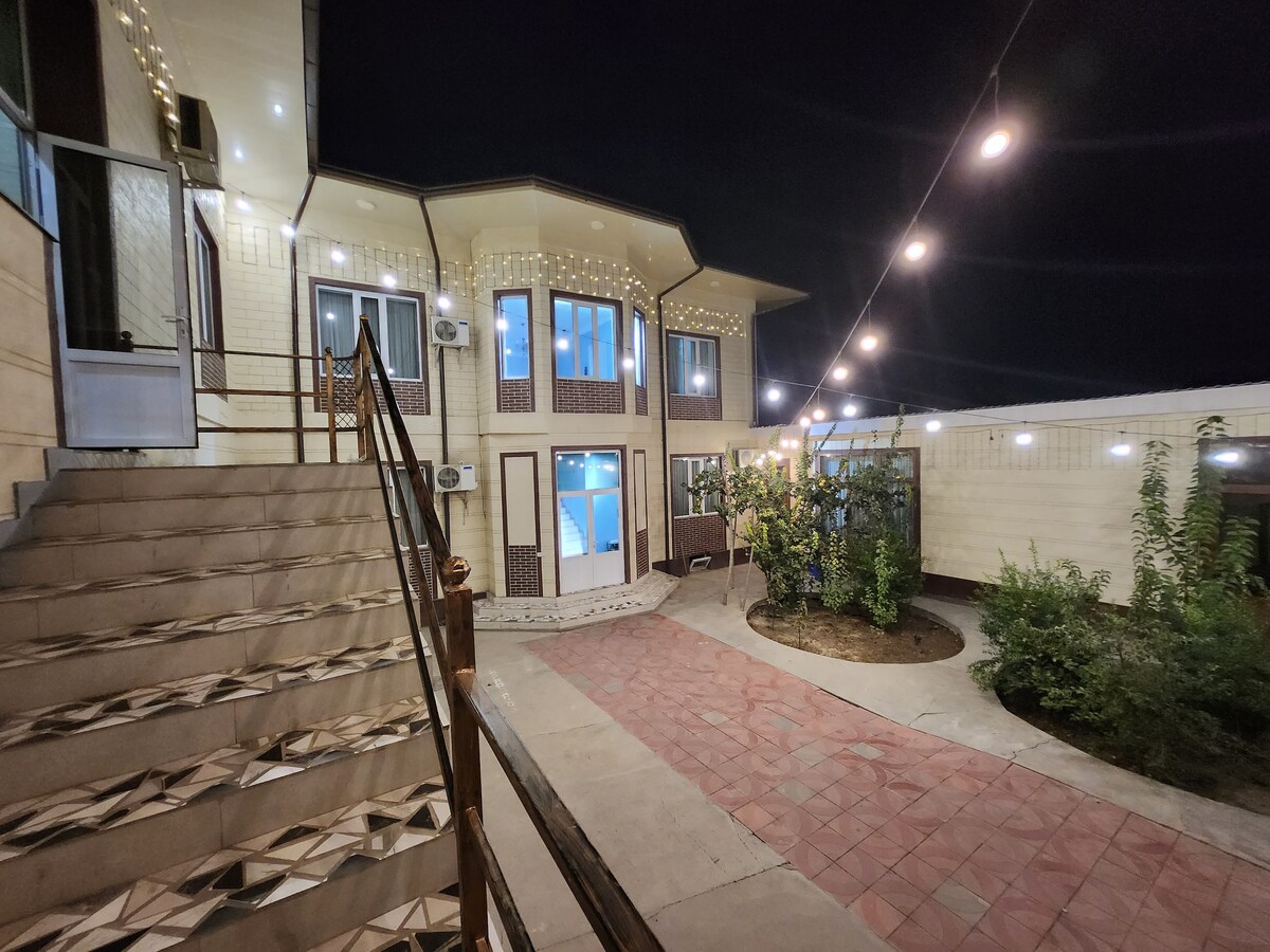 An outdoor area is illuminated by soft string lights. A spacious staircase leads to an entrance, with landscaped greenery visible in the courtyard. The two-story building features multiple windows, allowing light to spill into the surrounding space.