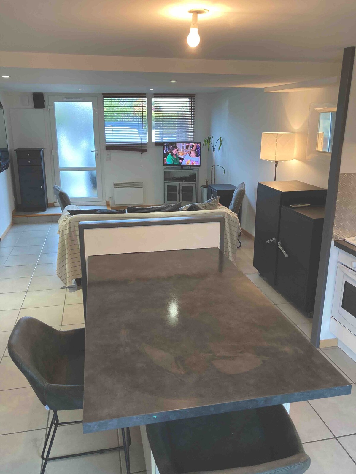 The interior space features an open-plan design with a dining area highlighted by a modern rectangular table and two dark chairs. A cozy living area is visible in the background, showcasing a small TV and a lamp providing warm light. Large windows allow natural light to fill the room.