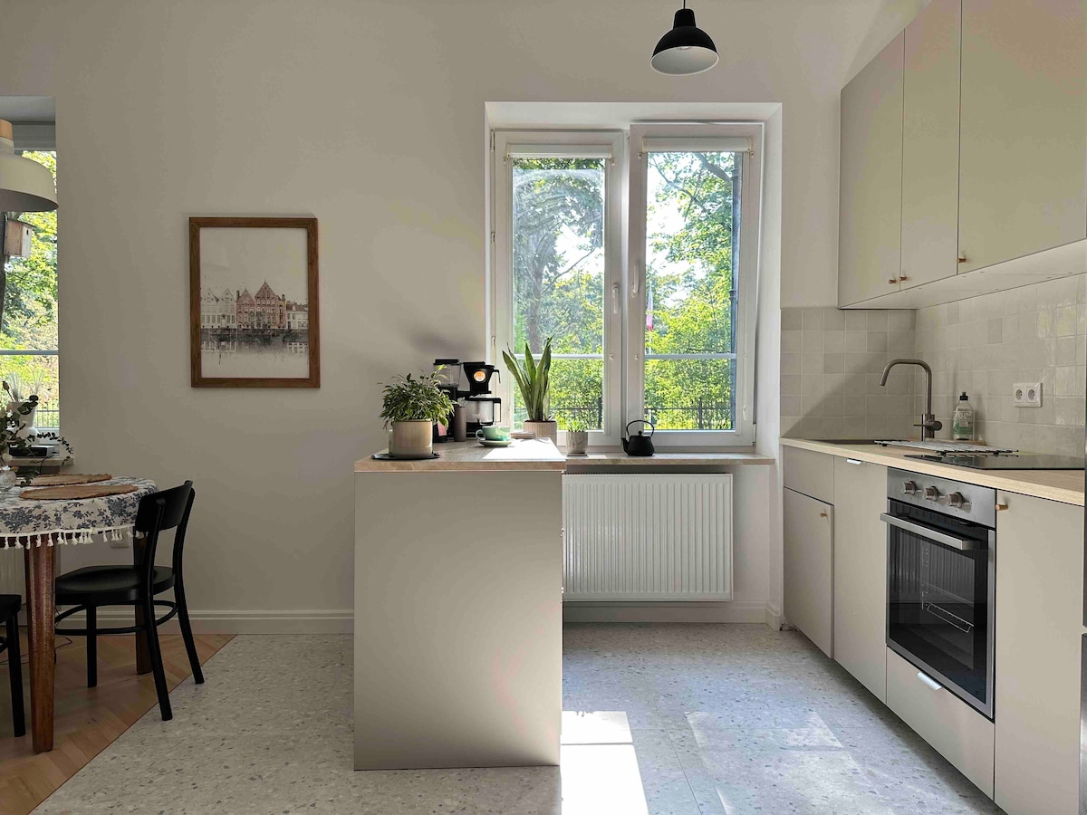 A well-lit kitchen area features a modern design with a central island and essential appliances, including an oven and a coffee maker. Natural light enters through large windows, illuminating a small potted plant. A dining space is visible in the background.