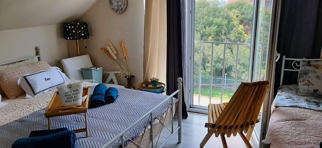 A bright bedroom features a spacious bed with neutral linens and decorative pillows. A small side table with books is positioned next to the bed. A wooden folding chair offers additional seating, while large glass doors provide access to a balcony overlooking greenery.